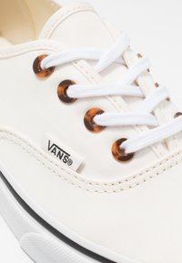 Canvas-Sneaker in Creme mit weißen Schnürsenkeln, braunen Ösen, schwarzem Sohlenrand und einem Vans-Logo-Label an der Seite.
