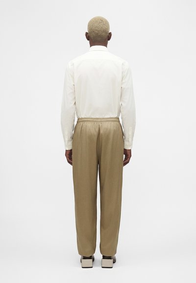 Άνδρας με κοντά ξανθά μαλλιά, φορώντας λευκό μακρυμάνικο πουκάμισο, μπεζ παντελόνι με ελαστική μέση και μπεζ παπούτσια με ξεχωριστή φιάπα, με πλάτη.