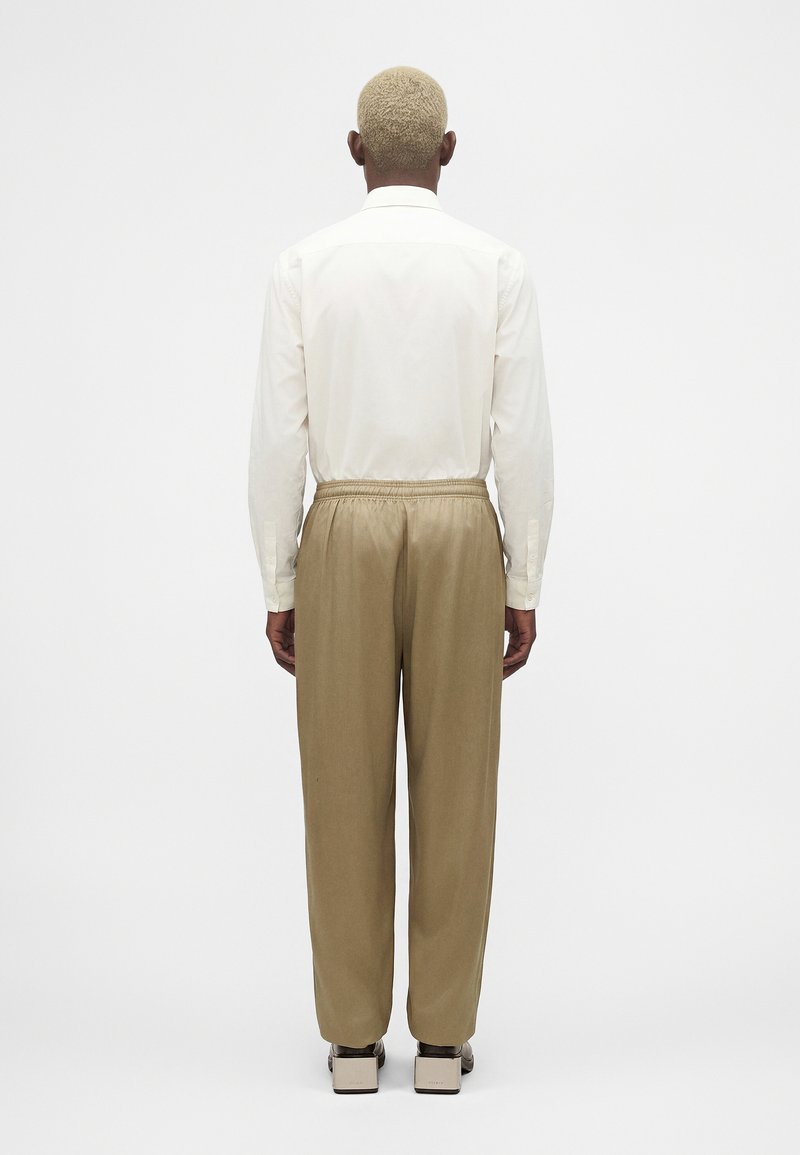 Homme aux cheveux blonds courts portant une chemise blanche à manches longues, un pantalon beige avec une ceinture élastique et des chaussures beiges à talons blocs, vu de dos.