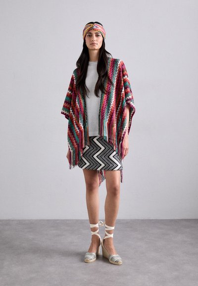 Bunt gestreifter Strickponcho mit Fransen; graues Oberteil; schwarz-weißer Chevron-Rock; gewebte silberne Keil-Espadrilles mit Schnürsenkeln.