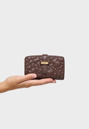 Mano sosteniendo una pequeña cartera de cuero marrón texturizado con relieve floral y un broche de metal dorado sobre un fondo gris claro.