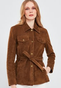 Bruine suède jas met een kraag, sluiting met knopen aan de voorkant, twee borstzakken en een tailleband voor een verstelbare pasvorm. Soepele textuur, tailormade ontwerp.