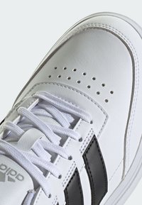 White leather sneaker featuring black side stripes, prominent perforations on the toe, and white laces. Smooth texture with contrasting accents.