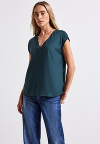 Maglietta corta teal con scollo a V, tessuto morbido, vestibilità rilassata e orlo leggermente arrotondato, abbinata a jeans azzurri chiari.