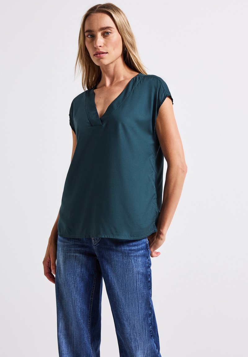 Maglietta corta teal con scollo a V, tessuto morbido, vestibilità rilassata e orlo leggermente arrotondato, abbinata a jeans azzurri chiari.