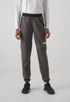 Person trägt dunkelgraue North Face Jogginghosen mit elastischem Bund und Bündchen, kombiniert mit schwarzen Schuhen und weißem Langarmshirt.