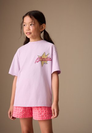Ung pige med mørkt hår i en hestehale, iført en lys lilla oversize T-shirt med "Blondie"-logo og teksturerede pink shorts.