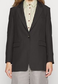 Musta bleiseri, jossa on yksi nappi, kaksi etutaskua ja lovetettu kaulus. Päällä raidallinen vihreä ja valkoinen paita. Yksinkertainen kankaan rakenne.