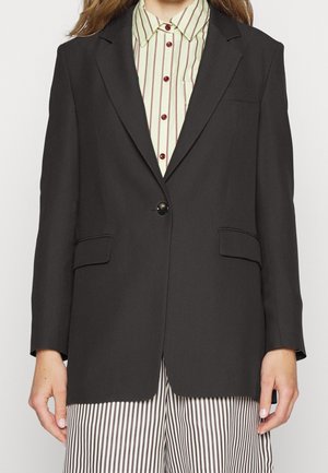 Zwarte blazer met één knoop, twee voorkruimels en een notch revers. Gedragen over een gestreept groen-wit shirt. Effen stofstructuur.