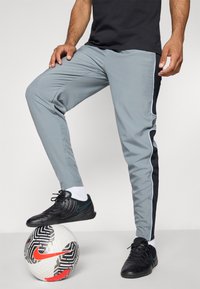 Grijze sportbroek met zwarte zijstreep, gecombineerd met zwarte voetbalschoenen op een gestippelde bal. Glad stof met een taps toelopende pasvorm.