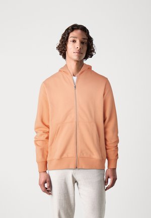 Felpa con zip color pesca realizzata in tessuto morbido, dotata di una tasca frontale, maniche lunghe e cappuccio con cordino. Abbinata a pantaloni chiari.
