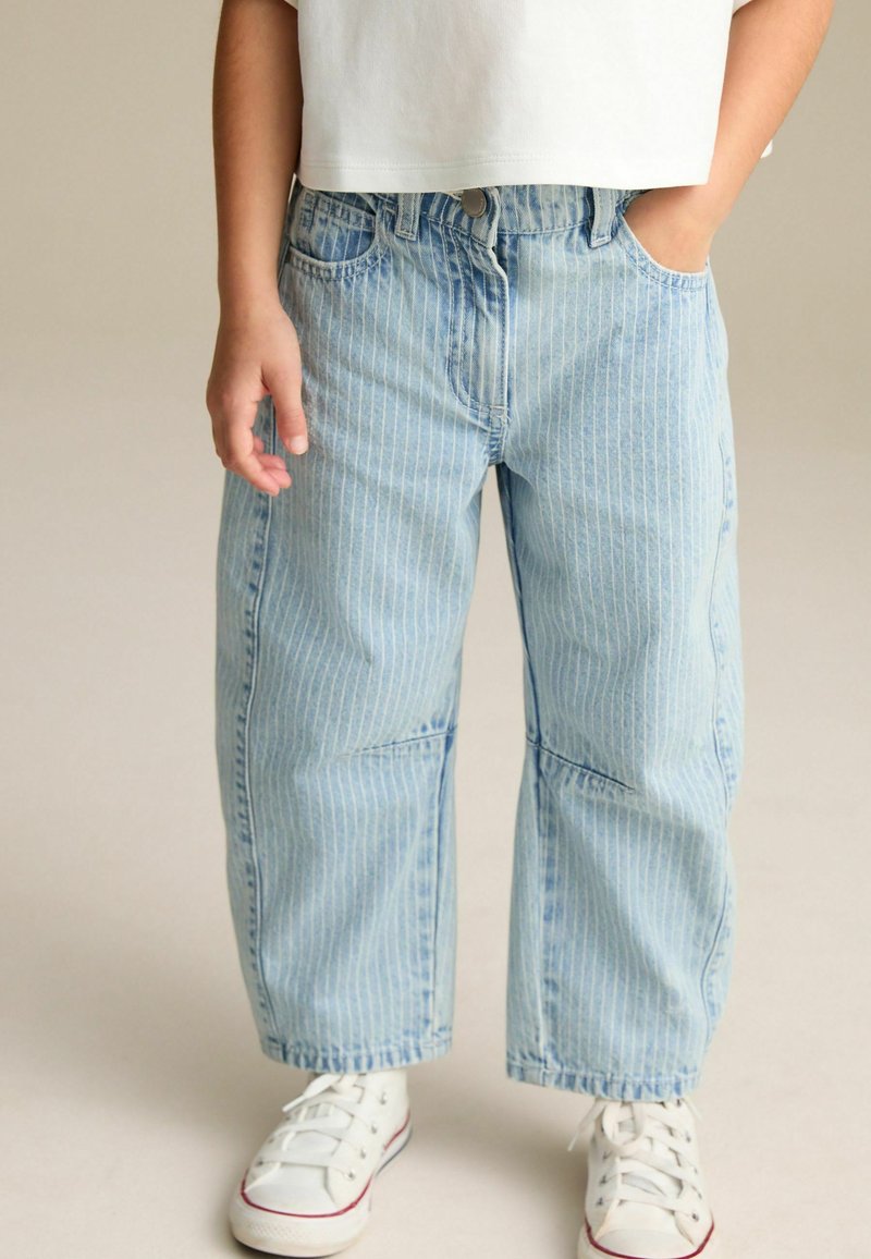 Jean en denim bleu clair avec des rayures blanches verticales, offrant une coupe loose, des poches avant et une taille à fermeture à bouton. Associé à des baskets blanches.
