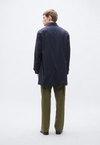 Tumesinine veekindel mantel kõrge krae, pikkade varrukatega ja lõdva lõikega, kombineeritud oliivroheliste kaubapükstega ning tumedate jalatsitega.