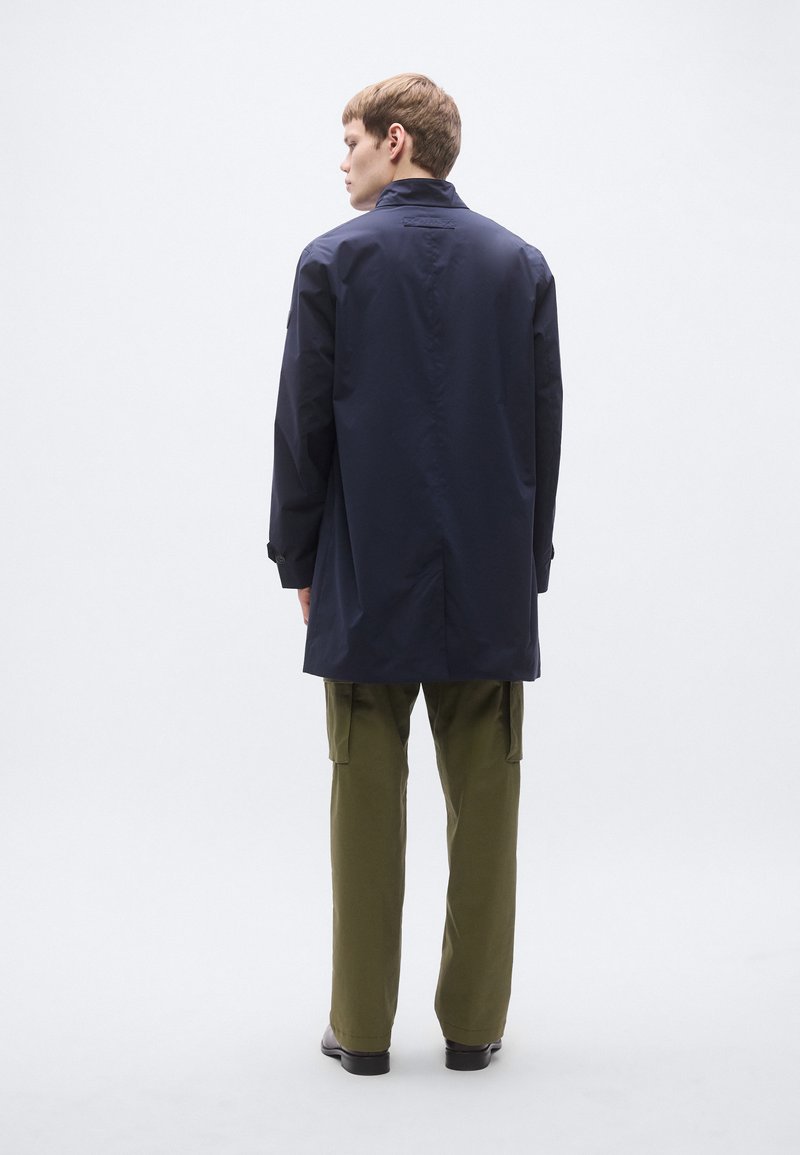 Navy waterproof coat with a high collar, long sleeves, and a relaxed fit, paired with olive green cargo pants and dark footwear.