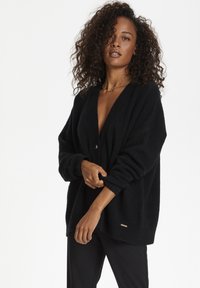 Black oversized cardigan with a deep V-neck, featuring a single button closure and ribbed cuffs. Soft texture and minimalist design.