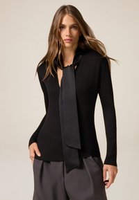 Maglietta nera a maniche lunghe con scollatura profonda e una cravatta in satin abbinata. Design aderente, struttura liscia, abbinata a pantaloni grigi scuri.