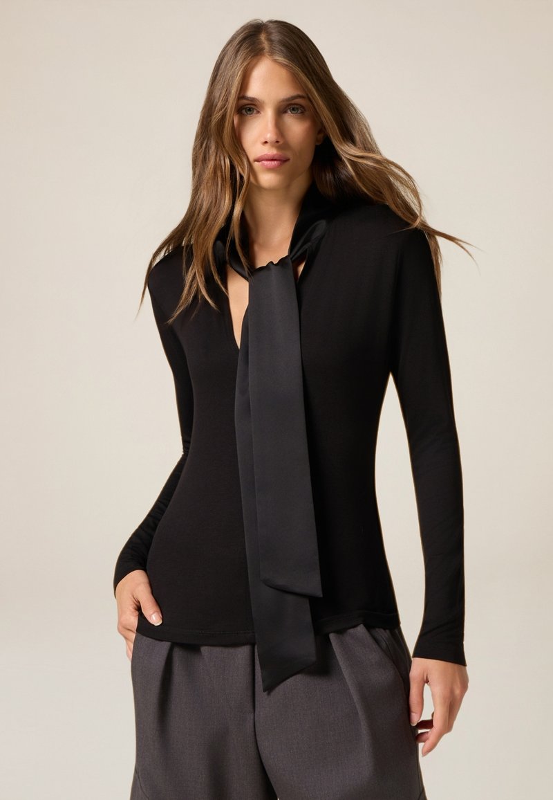 Maglietta nera a maniche lunghe con scollatura profonda e una cravatta in satin abbinata. Design aderente, struttura liscia, abbinata a pantaloni grigi scuri.