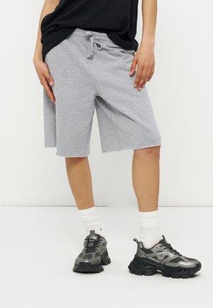 Osoba nosząca jasnoszare krótkie spodenki do kolan z troczkiem, białe skarpetki, metalicznie szare masywne sneakersy oraz częściowo widoczna czarna koszulka.