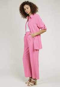 Chemise en lin rose et pantalon large à coupe décontractée avec une fermeture à boutons, associés à des sandales à talons beiges avec bride autour de la cheville.