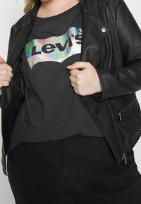 Czarna skórzana kurtka noszona na ciemnoszarym T-shircie z holograficznym logo Levi's. Całość dopełniają czarne spodnie.