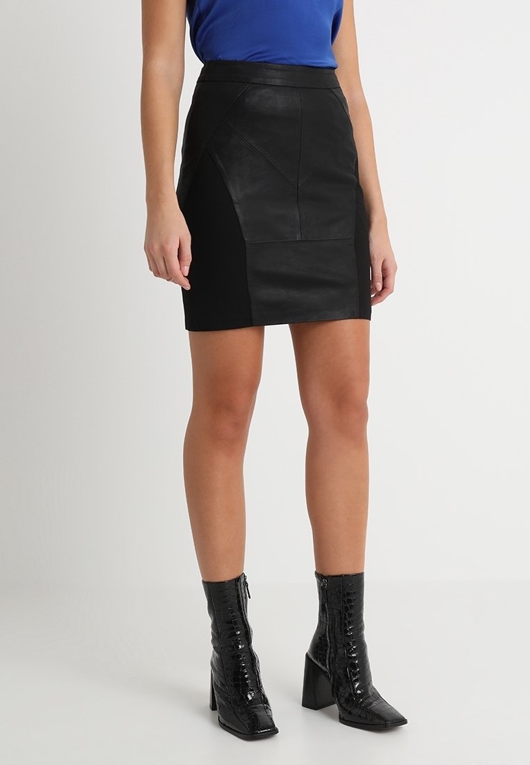 Musta nahkainen minihame, jossa on teksturoituja yksityiskohtia ja istuva malli, yhdistetty kiiltäviin mustiin nilkkureihin, joissa on krokotiilikuosi.