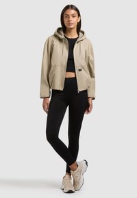 Femme portant une veste à capuche beige sur un crop top noir et un legging, debout jambes croisées, baskets beiges sur fond blanc.