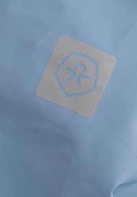 Ljusblått tyg har en fyrkantig silverlogotyp med en stiliserad människ Gestalt, centrerad på materialets släta textur.