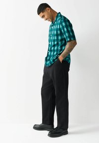 Cămașă cu mânecă scurtă de culoare teal și negru, cu carouri și nasturi, asociată cu pantaloni negri și pantofi negri, având o croială lejeră și o textură netedă.