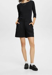 Czarne szorty z fałdami, bocznymi kieszeniami i elastycznym paskiem. Noszone z dopasowaną czarną bluzką i czarnymi loafersami. Widoczne białe skarpetki kostki.