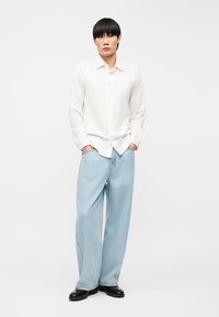 Νέος άνδρας στέκεται με τα χέρια στις τσέπες, φοράει λευκό πουκάμισο με μακριά μανίκια, ανοιχτό μπλε τζιν με φαρδιά μπατζάκια και μαύρα παπούτσια σε λευκό φόντο.