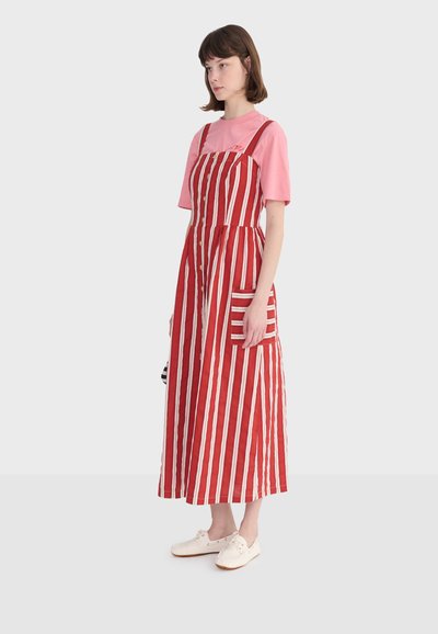 Jeune femme aux cheveux châtains courts portant un t-shirt rose sous une robe sans manches rouge et blanche rayée avec des boutons et des chaussures blanches, debout.