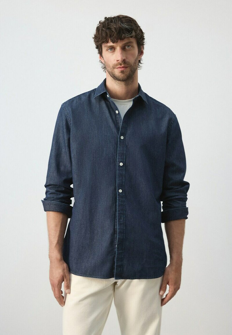 Homme aux cheveux bruns portant une chemise en jean bleu foncé sur un t-shirt blanc et un pantalon beige clair, debout devant un fond clair uni.