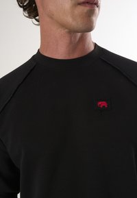 Must must, mille ümmarguse kaeluse ja raglaansleevedega. Vasakul rinnal on silmapaistev punane elevandi logo plekk. Sile tekstuur.