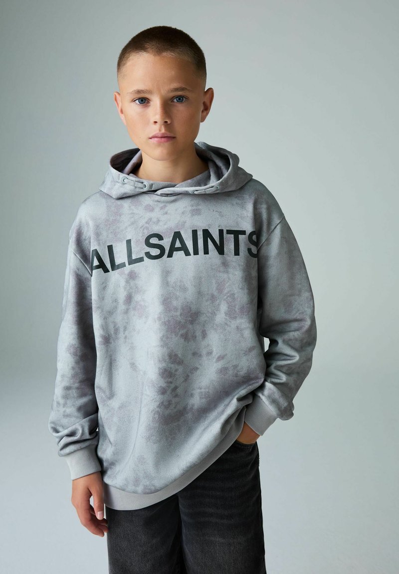 Šedá mikina s zavazovacím kapucím a klokaní kapsou, s černým logem "ALLSAINTS" na hrudi. Kombinováno s tmavými džíny.