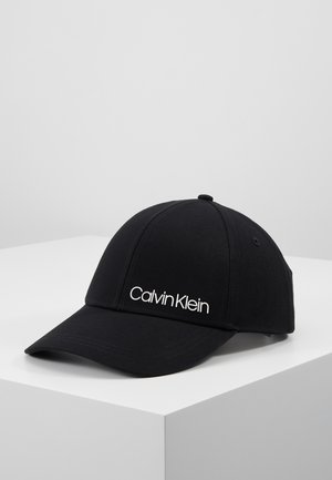 Black Calvin Klein baseball cap with curved brim placed on white surface against plain gray background.