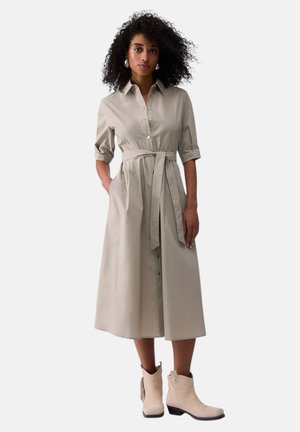 Donna con capelli ricci che indossa un vestito midi beige con bottoni e cintura in vita e stivaletti beige, in piedi davanti a uno sfondo semplice.