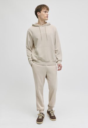 Joven con sudadera beige con bolsillo frontal y pantalones deportivos a juego, de pie y mirando hacia un lado contra un fondo claro y sencillo.