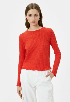 Suéter de punto acanalado rojo con cuello redondo, mangas largas y dobladillo festoneado. Lleva puesto sobre pantalones cargo blancos, mostrando textura y ajuste.