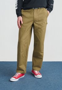 Άνδρας που φορά χακί παντελόνι εργασίας, μαύρη ζώνη, μαύρο μακρυμάνικο πουκάμισο και κόκκινα πάνινα παπούτσια, στέκεται σε μπλε χαλί.