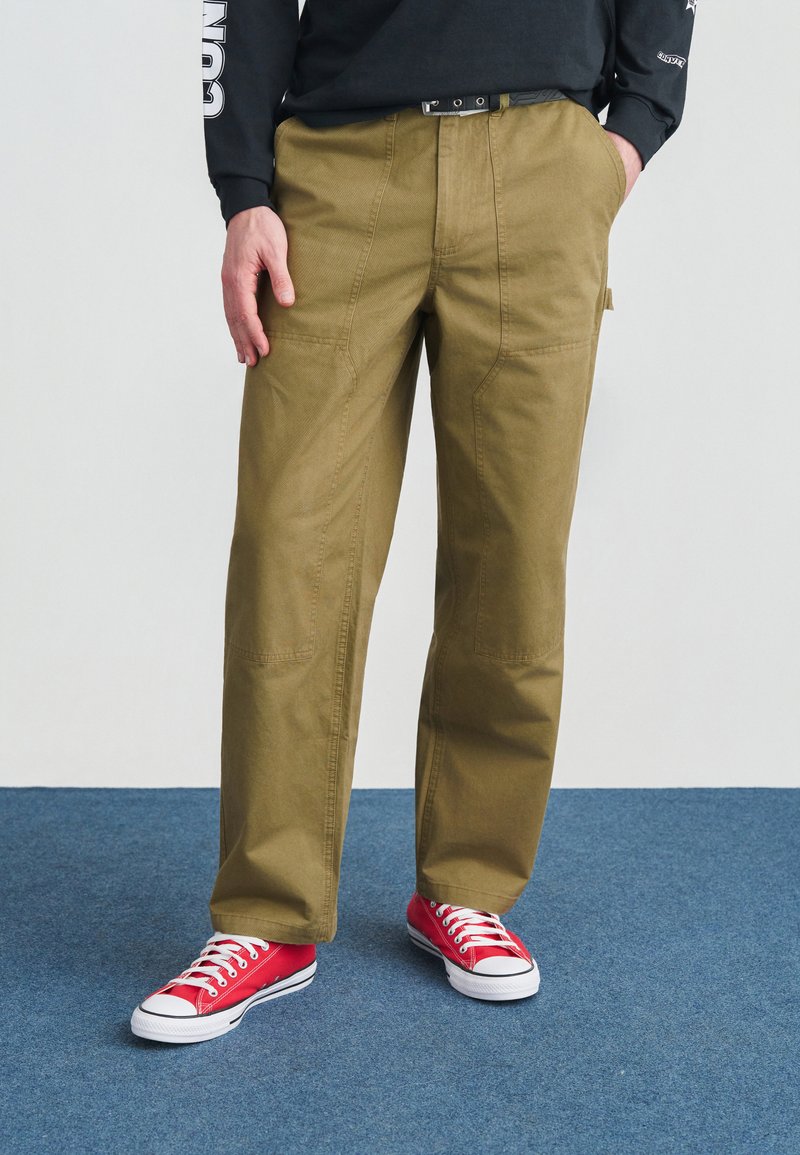 Άνδρας που φορά χακί παντελόνι εργασίας, μαύρη ζώνη, μαύρο μακρυμάνικο πουκάμισο και κόκκινα πάνινα παπούτσια, στέκεται σε μπλε χαλί.