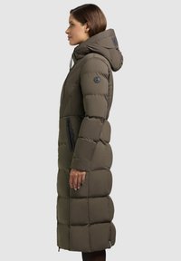 Lang, olivengrøn dunjakke med hætte, der har et quiltet design, sidelommer og et logo-tryk på kraven. Glat tekstur.