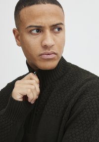 Dunkelgrüner Reißverschluss-Pullover mit strukturierten Strickmustern und gerippten Bündchen. Nahaufnahme, die eine Hand zeigt, die den Reißverschluss zieht.