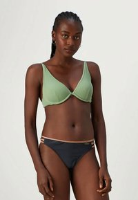 Grønn ribbestrikket bikini-oberdel med V-hals og svarte bikini-bunner med gull detaljerte stropper. Glatt tekstur, minimalistisk design.