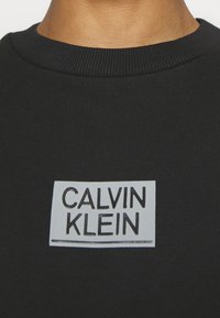 La felpa nera in cotone presenta un collo a costine e un'etichetta rettangolare grigia con la scritta "CALVIN KLEIN" in caratteri neri in grassetto.