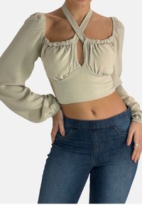 Hellbeige langärmliges Oberteil mit quadratischem Ausschnitt, gerafften Details und überkreuzten Trägern, kombiniert mit dunkelblauen, eng anliegenden Jeans.