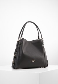 Musta nahkabag, jossa on löysä muotoilu, kaksi pyöreää kahvaa ja kultasävyiset yksityiskohdat. Edessä on pieni logo koriste.