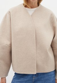 Šviesiai ruda vilnonė trumpa striukė su V formos iškirpte, dėvima ant baltos palaidinės, kartu su mėlynomis džinsinėmis kelnėmis.