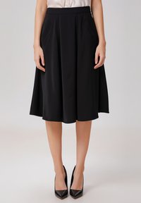 Jupe noire en A en tissu lisse, dotée de poches latérales et d'une taille ajustée, associée à des escarpins noirs pointus.