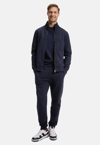 Veste zippée marine par-dessus un sweatshirt et un pantalon de jogging assortis. Modèle portant des baskets blanches et noires. Tissu doux avec des poignets et une taille élastiques.