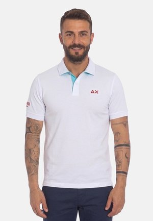 Uomo sorridente con capelli corti e barba, indossa una polo bianca con logo rosso e pantaloni blu, mostrando tatuaggi su entrambi gli avambracci.